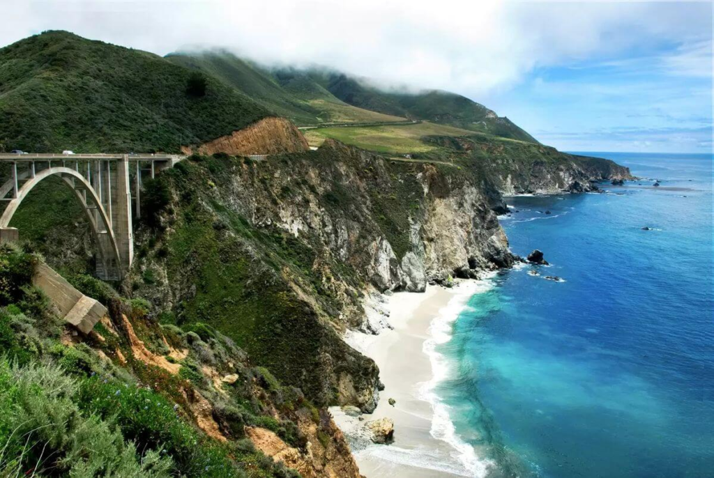 Big Sur coastline with rugged cliffs, turquoise ocean waves, and the iconic Bixby Creek Bridge along Highway 1.