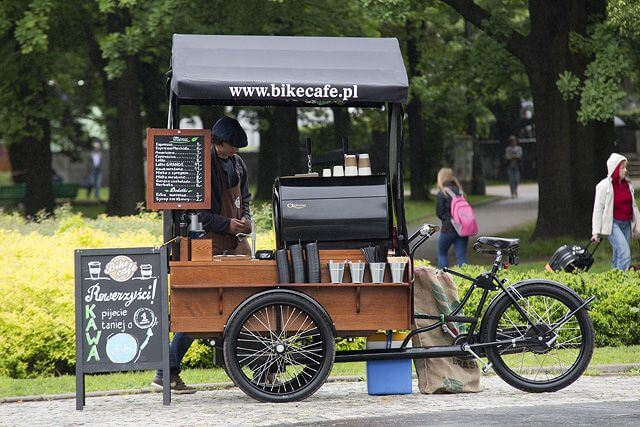 Coffee cart on a bike in a park, with a sign displaying the menu and a barista serving drinks