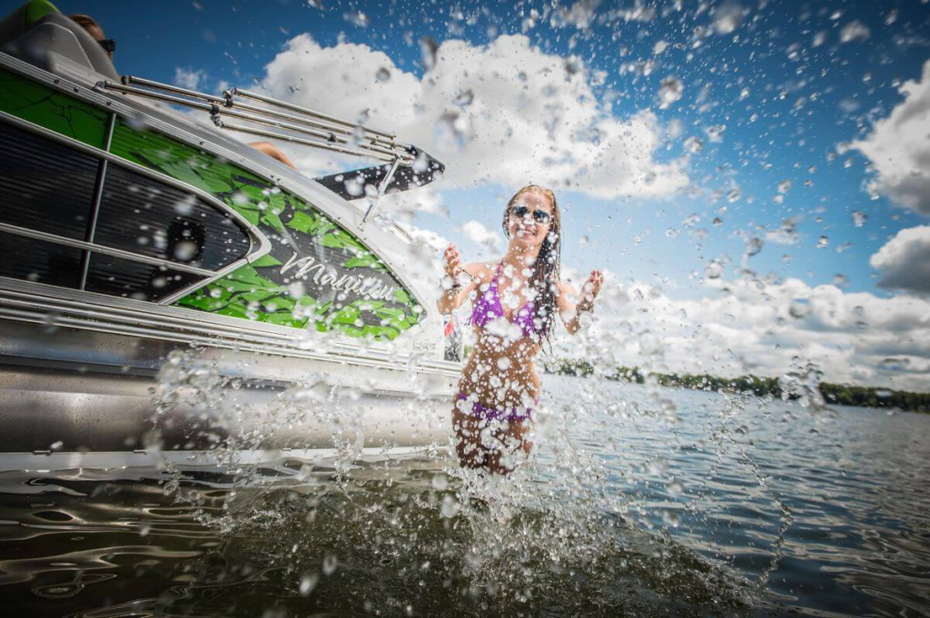 Girl in a bikini splashing water next to a green and silver pontoon boat on a sunny day.