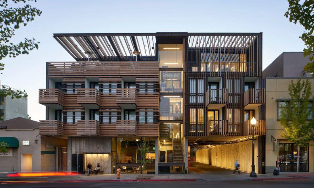 Modern wood-accented hotel building with balconies, open-air design, and warm evening lighting in a downtown setting.