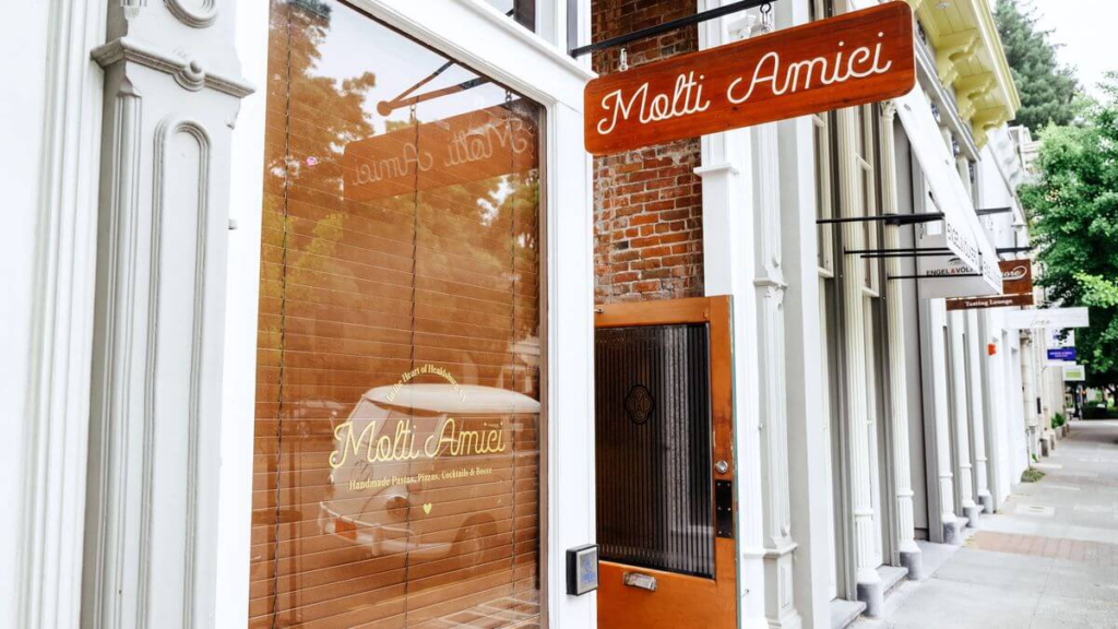 Exterior of Molti Amici restaurant with wooden signage, brick walls, and large front window on a quiet street.