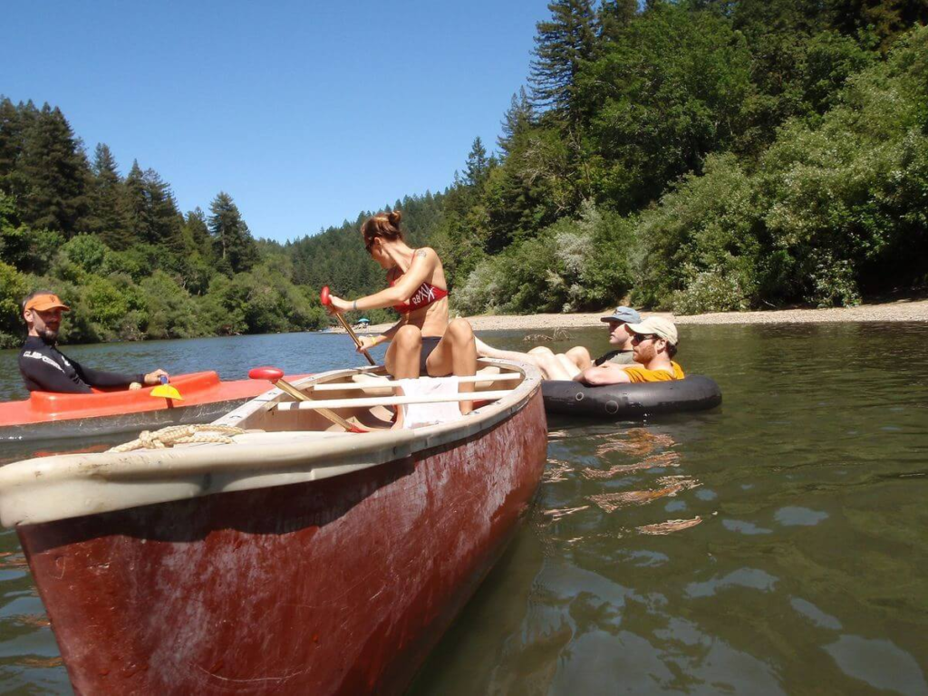 "People kayaking and tubing on the Russian River surrounded by trees on a sunny day."