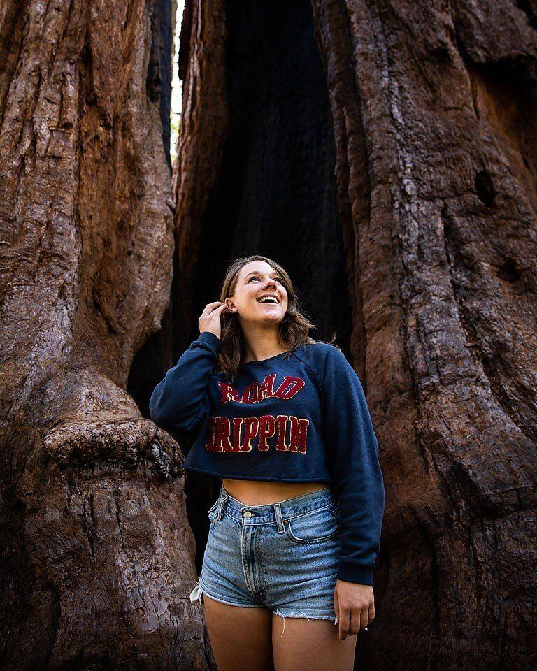 "Smiling woman standing in front of a giant redwood tree, wearing a 'Road Trippin' sweatshirt."