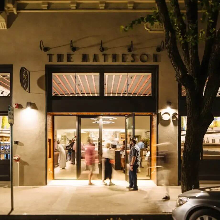 Nighttime exterior of The Matheson restaurant with warm lighting and guests entering through glass doors.