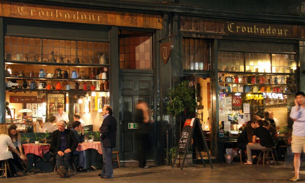 Nighttime street view of the Troubadour café with vintage decor, people dining outdoors, and warm glowing lights.