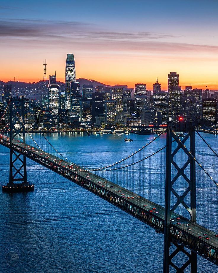 "San Francisco city skyline with the Bay Bridge at twilight, showcasing lights and reflections over water."
