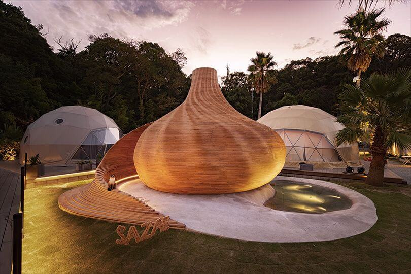 Unique wooden dome structure at Sana Mane glamping site in Naoshima, Japan, surrounded by greenery and modern geodesic tents at sunset.