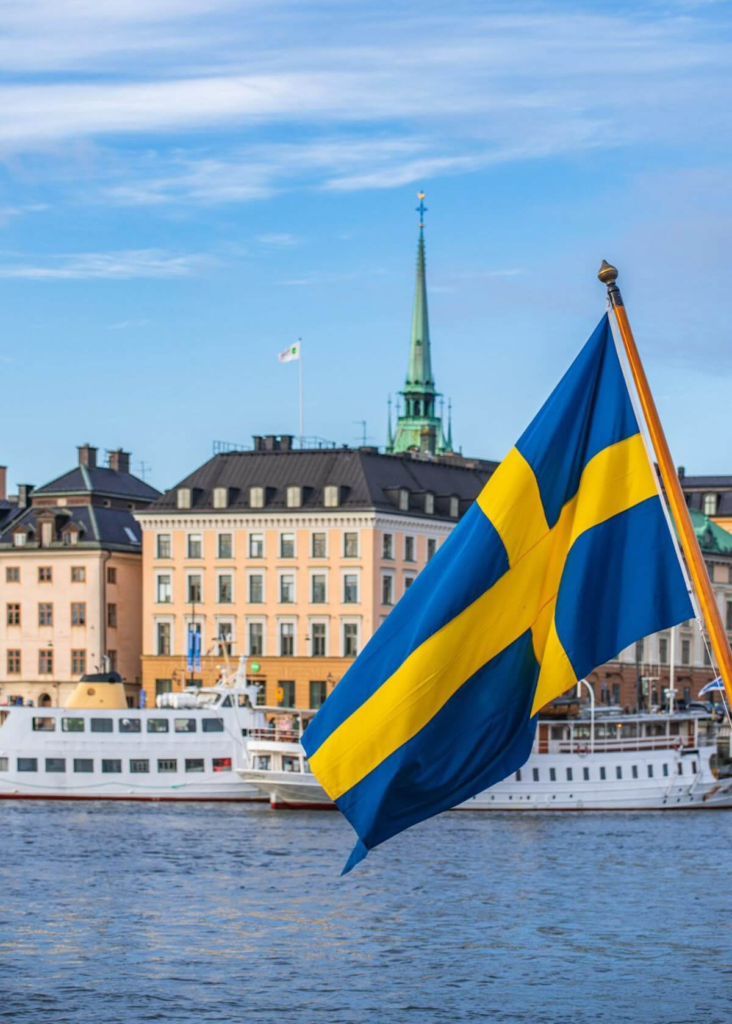 Swedish flag waving over Stockholm’s waterfront with historic buildings and boats in the background.
