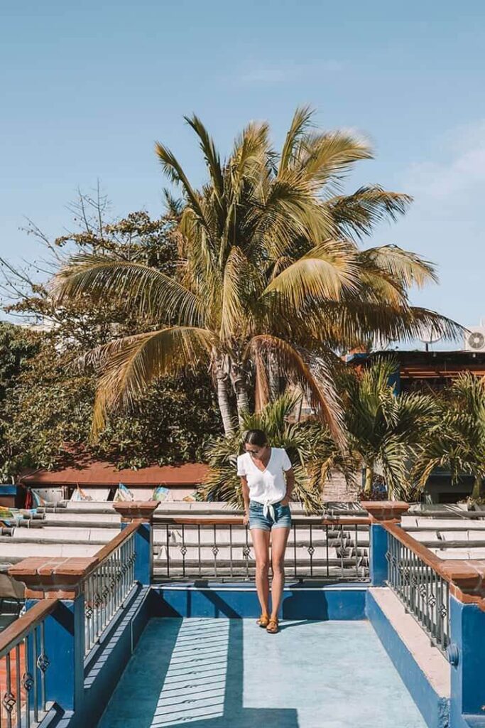 "Young woman in shorts standing on a blue balcony with palm trees and bright sunlight in Todos Santos, Mexico."